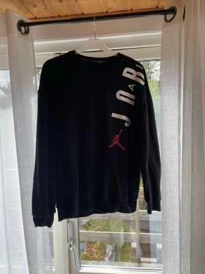 Svart sweatshirt från Air Jordan - Svart sweatshirt från Air Jordan med vit text och den klassiska röda Jumpman-loggan på framsidan. Tröjan har rund halsringning och ribbade muddar vid ärmslut och nederkant. Perfekt för en sportig och avslappnad stil.