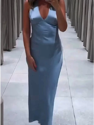 Ljusblå långklänning i satin - Elegant ljusblå långklänning i glansigt satinmaterial med v-ringad halsringning. Klänningen har en smickrande passform och faller mjukt längs kroppen, perfekt för speciella tillfällen.