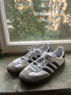 Adidas Samba vita och svarta sneakers - Klassiska Adidas Samba sneakers i vitt och svart med grå mockadetaljer på tån och mörkbrun sula. Ovandelen är i läder och mocka. Ett par skor som funkar till allt! De är i super bra skick, i princip aldrig använda!