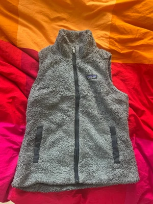 Grå fluffig pälsväst med dragkedja - Mysig grå pälsväst från Patagonia med hög krage och hel dragkedja framtill. Västen har två fickor med svarta detaljer och är gjord i ett fluffigt, mjukt material som ger en avslappnad och trendig look.