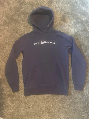 Marinblå hoodie från Sail Racing - Säljer en marinblå hoodie från Sail Racing med vit logga på bröstet. Klassisk modell med huva och magficka framtill. Tröjan är i jätte bra skick👍Tillverkad i mjukt material som känns skönt mot huden. Perfekt för dig som gillar stilren och sportig look. Grym nu mot hösten och de kyligare dagarna❄️ Fraktar samma dag plagget är köpt❤️‍🩹 