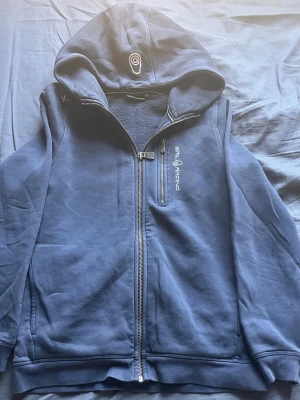 Marinblå hoodie från Sail Racing - Säljer en marinblå hoodie från Sail Racing med dragkedja framtill och broderad logga på bröstet. Hoodien har en stor huva och mjukt fleecefoder på insidan. Perfekt för dig som gillar sportig och avslappnad stil.