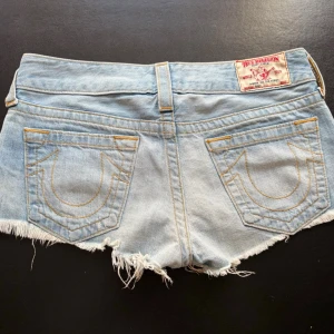 True Religion Jeansshorts - Säljer ett par korta ljusblå jeansshorts från True Religion med fransig, rå kant och snygga bakfickor. Shortsen har mycket låg midja, knäpps med knappar samt slitna detaljer.