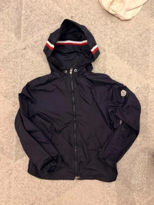 Äkta moncler vindjacka - Snygg mörkblå vindjacka med huva och dragkedja framtill. Huvan har röda och vita ränder och jackan har ett emblem på ärmen. Tillverkad i lätt och tåligt material, perfekt för blåsiga dagar.