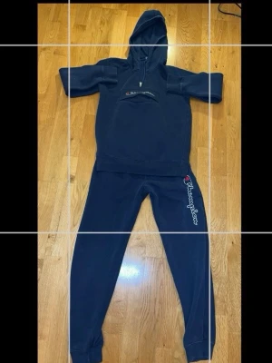 Mörkblå Champion loungewear-set - Säljer ett mörkblått loungewear-set från Champion med huva och dragkedja på tröjan samt matchande byxor med logga längs benet. Setet är i mjukt material och har ribbade muddar vid ärmar och benslut. Perfekt för chill dagar hemma.