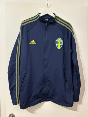 Blå Sverige träningsjacka Adidas - Mörkblå träningsjacka från Adidas med Sveriges landslagsemblem på bröstet och gula detaljer. Jackan har dragkedja, två fickor och klassiska Adidas-ränder längs ärmarna. Perfekt för träning eller att bära till vardags.