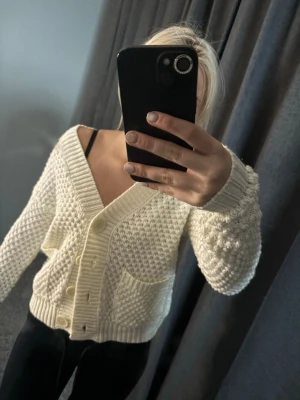 Vit stickad kofta från Vero Moda - Säljer en krämvit stickad kofta från Vero Moda i storlek XS. Koftan har grovstickat mönster, djup V-ringning, stora knappar framtill och två fickor. Perfekt för lager på lager och riktigt mysig att bära till jeans eller kjol.
