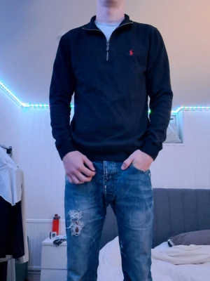 Svart halvzip tröja från Polo Ralph Lauren - Svart långärmad tröja från Polo Ralph Lauren med halv dragkedja och röd broderad logga på bröstet. Tröjan har ribbade muddar och hög krage, perfekt för en clean och stilren look.