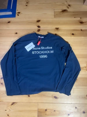 Svart långärmad topp Acne Studios - Cool blå långärmad topp från Acne Studios med tryck 'Acne Studios STOCKHOLM 1996' på bröstet. Tillverkad i mjuk bomull och har en relaxed fit. Perfekt för dig som gillar minimalistisk och stilren design.