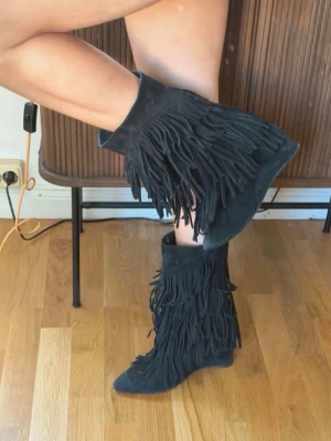 skor med fransar - Säljer ett par svarta cowboy boots i mocka med flera lager långa fransar längs skaftet. Bootsens tå är spetsig och klacken är låg och bred. Perfekta för dig som vill sticka ut med en cool och trendig look.