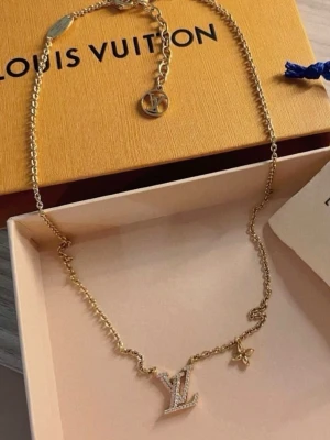 Guldhalsband med diamanter intryckta - Snyggt halsband i guldton med en liten hint av rosa underton med en glittrande logga dekorerad med små stenar och en liten blomformad detalj. Kedjan är tunn och elegant, perfekt för att lyfta vilken outfit som helst. Ask följer inte med (har inte den)