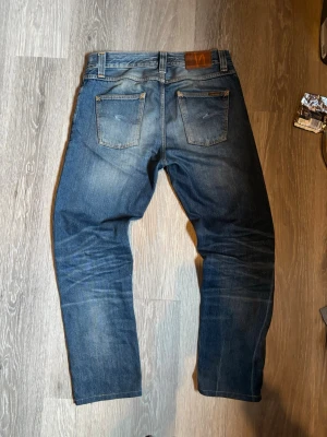 Nudie Jeans | Straight fit - Säljer ett par klassiska blå jeans från Nudie Jeans med snygg slitning och raka ben. Jeansen har två bakfickor med vågigt broderi och en brun läderpatch i midjan. Perfekta för en avslappnad och trendig stil. Storleken är 30/34 men är uppsydda till 30/32 för att vara perfekta