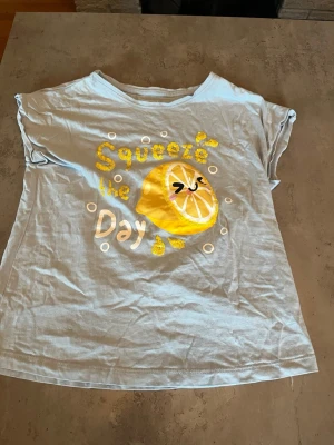 Ljusblå t-shirt med citrontryck Firefly - Ljusblå t-shirt från Firefly med korta ärmar och ett färgglatt citrontryck samt texten 'Squeeze the day' i gult och vitt framtill. Tillverkad i mjuk bomull, perfekt för en avslappnad och lekfull stil.