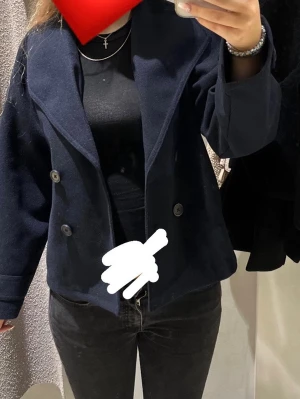 Mörkblå kort kappa - Snygg mörkblå kort kappa med dubbelknäppning och bred krage. Rocken har en rak passform och är tillverkad i ett ull-liknande material. Perfekt för dig som vill ha en stilren och enkel look till hösten eller vintern.