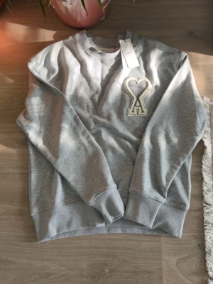 Grå sweatshirt från Ami Paris - Säljer en stilren grå sweatshirt från Ami Paris med det ikoniska hjärta-A-märket broderat på bröstet. Tröjan har rund halsringning, ribbade muddar och är gjord i mjukt bomullsmaterial. Perfekt för en avslappnad och trendig look.