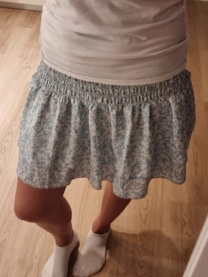 Blommig kortkjol med volang - Söt kortkjol med ljusblått och vitt blommönster. Kjolen har bred resår i midjan och volangkant nertill som ger en luftig och somrig känsla. Perfekt för dig som gillar en feminin och avslappnad stil.
