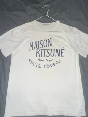 Maison Kitsuné vit t-shirt Paris - Vit t-shirt från Maison Kitsuné med tryck i marinblått framtill där det står 'Maison Kitsuné Palais Royal Paris, France'. Klassisk rund hals och korta ärmar. Tillverkad i mjuk bomull för en skön och avslappnad stil. Lite små fläckar vid armarna skriv om ni vill ha bild på dom