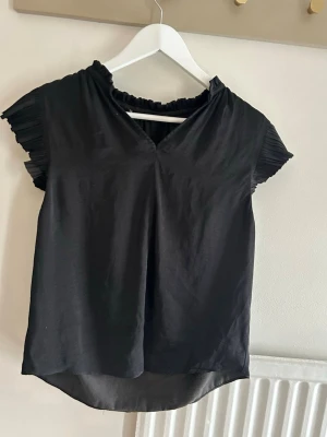 Svart topp med plisserade ärmar - Snygg svart topp med korta plisserade ärmar och rynkad halsringning. Toppen har en lätt v-ringning framtill och är tillverkad i ett tunt, mjukt material som faller fint. Perfekt att styla med jeans eller kjol för en trendig look. I silkes material 