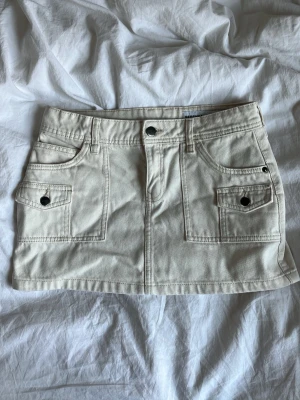 Beige kort jeanskjol med fickor - Snygg beige kortkjol i jeansmaterial med flera fickor fram och bak samt bälteshällor från Bershka. Kjolen har knapp och dragkedja framtill och coola detaljer med nitar på fickorna