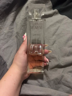 Eternity Moment parfym - Elegant parfymflaska i genomskinligt glas med rektangulär form och stilren design. Flaskan har texten 'Eternity Moment' tryckt framtill och ett transparent lock. Innehållet är ljusrosa och flaskan känns lyxig och modern.