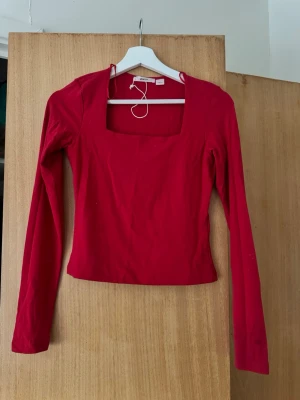 Röd långärmad topp med fyrkantig ringning - Snygg röd topp med lång ärm och fyrkantig ringning. Modellen är figurnära och passar perfekt till jeans eller kjol. Tillverkad i mjukt material som känns skönt mot huden. Enkel men trendig design som lyfter vilken outfit som helst.