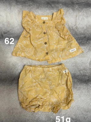 Gult blommigt set med volanger - Sött tvådelat set i ljusgul färg med vita blommönster. Toppen har volangärmar och knappar framtill, medan shortsen har resår i midja och benslut för bekväm passform. Perfekt för varma dagar och ger en gullig look.🌸 Fint använt skick. Storlek 62