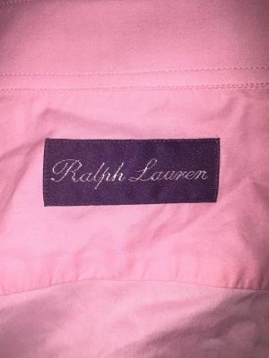 Rosa skjorta från PURPLE LABEL Ralph Lauren - Säljer en klassisk rosa skjorta från vintage PURPLE LABEL Ralph Lauren med lång ärm och knappar framtill. Skjortan har den ikoniska broderade loggan med polospelare på bröstet och är tillverkad i mjuk bomull. Perfekt för en stilren och avslappnad look. Skjortan är förmodligen gjord på 1990-2000 tal