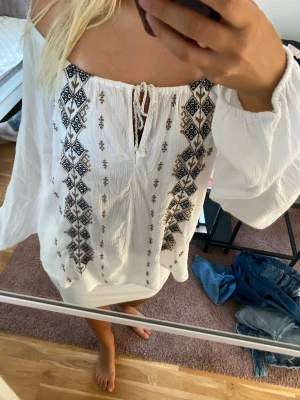 Vit offshoulder blus med brodyr - Säljer en vit offshoulder blus med svart och brun brodyr i boho-stil. Blusen har långa, vida ärmar och knytning framtill vid halsen. Materialet är tunt och luftigt, perfekt för sommardagar eller festivaler.