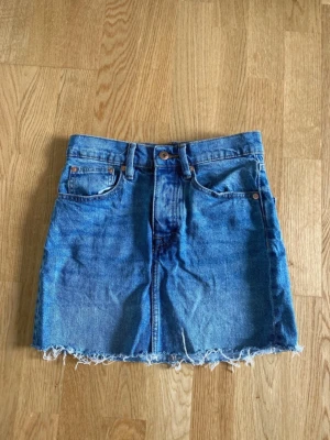 Blå jeanskjol  - Säljer en klassisk blå jeanskjol i kort modell med rå, fransig kant nedtill. Kjolen har fem fickor, bälteshällor och knappgylf. Perfekt till sommaren och enkel att styla med olika toppar.