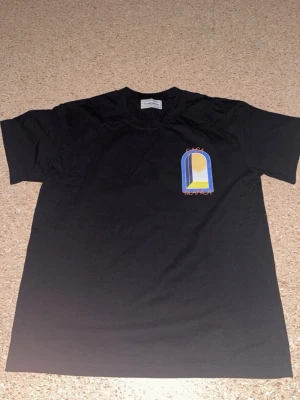 Svart Casa Blanca t-shirt med tryck - Svart t-shirt från Casa Blanca med färgglatt grafiskt tryck av ett fönster i blått, gult och vitt på både bröstet och ryggen. T-shirten har rund hals och korta ärmar. Perfekt för dig som gillar streetwear och unika prints.