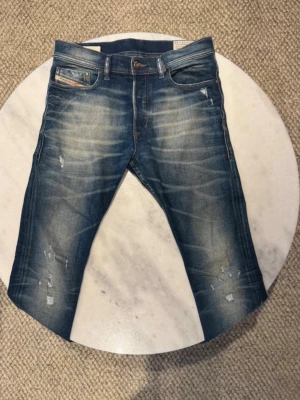 Blå slitna jeans med tvättade detaljer - Säljer ett par blå jeans med snygga slitningar och tvättade detaljer. Jeansen har fem fickor, raka ben och klassisk knappgylf. Materialet är denim i bomull och passformen är modern och avslappnad. Perfekta för en trendig och avslappnad stil.