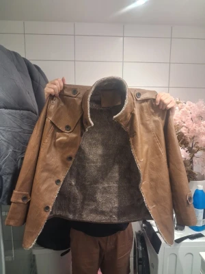 Brun skinnjacka med fuskpälsfoder - Snygg brun skinnjacka med stora knappar och två bröstfickor med lock. Jackan har ett mjukt, lurvigt foder på insidan som håller dig varm. Klassisk krage och raka ärmar med knappdetaljer vid ärmslut. Perfekt för dig som vill ha en retro och cool look.