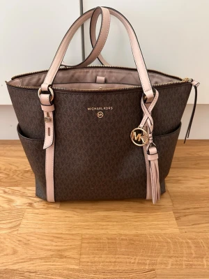 Brun handväska från Michael Kors - Snygg brun handväska från Michael Kors med ljusrosa detaljer och guldfärgade metalldetaljer. Väskan har två handtag, dekorativ tofs och MK-hänge, samt flera innerfack och dragkedja upptill. Tillverkad i tåligt konstläder med mönstrad yta.