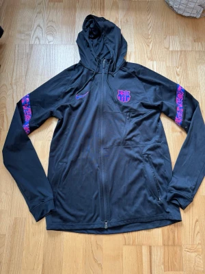 Svart Nike FC Barcelona tracksuit  - Säljer ett svart träningsset från Nike och FC Barcelona. Setet består av en hoodie med dragkedja och matchande byxor, båda med lila och rosa detaljer samt klubbmärke och Nike-logga. Materialet är mjukt och stretchigt, perfekt för träning eller chill.