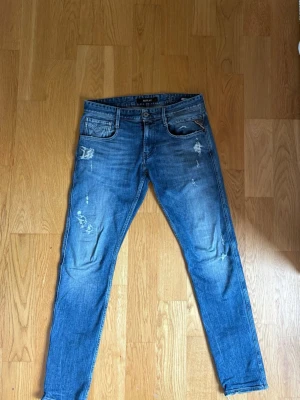 Blå slitna jeans från Replay - Säljer ett par blå jeans från Replay i storlek 30. Jeansen har en slim fit-modell med slitningar och detaljerade revor på benen för en trendig look. Klassisk femficksdesign och knappgylf. Perfekta för dig som gillar en avslappnad men stilsäker stil.