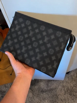 Svart monogram clutch - Snygg svart clutch  med klassiskt monogrammönster över hela väskan. Tillverkad i canvas med detaljer i svart läder och silverfärgad dragkedja . Perfekt storlek för det viktigaste och enkel att bära i handen.