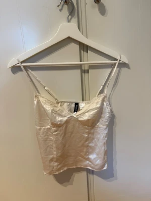 Champagnefärgad satin linnetopp - Snygg champagnefärgad linnetopp i satin med tunna axelband och spetsdetalj vid urringningen. Perfekt att styla med jeans eller kjol för en trendig look. Toppen har en lätt glansig finish och smickrande passform.