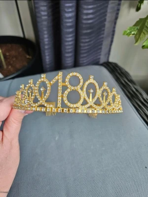 Guldigt diadem med siffran 18 - Snyggt diadem i guld med siffran 18 framtill, täckt av glittrande strass. Perfekt accessoar till 18-årsdagen för att sticka ut och känna sig extra speciell. Diademet har en klassisk tiaraform och är gjort av metall med gnistrande stenar.