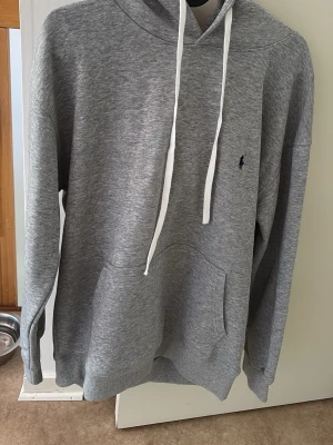 Grå raffe hoodie med huva och ficka - Snygg grå hoodie med huva och vita dragsnören. Klassisk känguruficka framtill och diskret broderad logga på bröstet. Mjuk och bekväm bomullsblandning, perfekt för chill dagar eller när du vill ha en clean look.