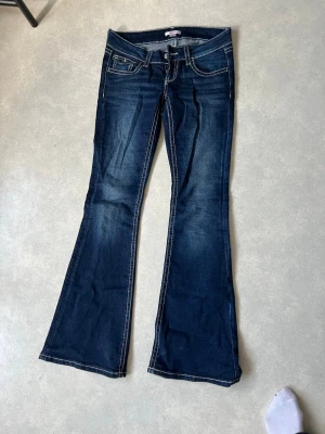 Mörkblå bootcut jeans med broderi - Säljer ett par mörkblå bootcut jeans med kontrastsömmar och snygga broderade detaljer på bakfickorna. Jeansen har låg midja och klassisk femficksdesign. Perfekta för dig som gillar en retro och avslappnad stil.