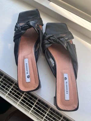 Svarta sandaler med knut från Blankens - Stilrena svarta sandaler från Blankens med fyrkantig tå och låg klack. Ovandelen har en snygg knutdetalj i mjukt läder och innersulan är beige. Perfekta för dig som gillar trendiga och bekväma skor med en twist. Modellen heter The Kajsa.
