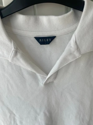 Vit piké t-shirt från Riley - Klassisk vit piké t-shirt från Riley med krage och v-ringad öppning. Tillverkad i mjuk bomull som känns skön mot huden. Enkel och stilren design som passar till det mesta. Perfekt för dig som gillar en clean och fräsch look.