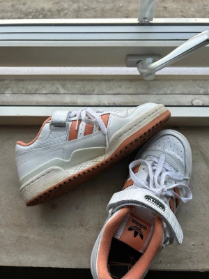 Adidas vita och orange sneakers - Snygga Adidas sneakers i vitt och orange med klassiska tre ränder på sidan. Skorna har kardborreband över snörningen och en gummisula i brunt. Ovandelen är i läder och textil, med vadderad häl och plös för extra komfort. Perfekta för en sportig och trendig look.