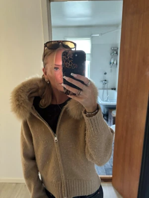 Beige pälsjacka med luva från Alessa - Säljer en beige pälsjacka med dragkedja och stor fluffig luva med pälsdetaljer från Alessa. Jackan har ribbade muddar och en avslappnad passform. Perfekt för kalla vinterdagar och ger en riktigt cozy vibe.