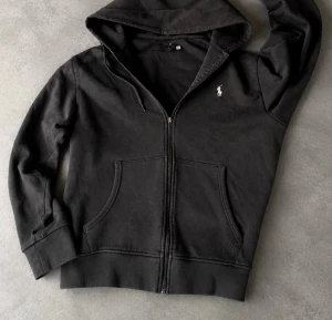 Svart Ralph lauren hoodie - Svart hoodie med dragkedja och huva. Klassisk design med fickor framtill och justerbara snören i huvan. Liten vit logga broderad på bröstet. Perfekt för en avslappnad och stilren look. Dragkedja salen man drar i har gått av