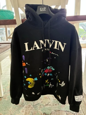 Svart hoodie LANVIN x DEPT. de la GALERIE - Säljer en svart hoodie från LANVIN x DEPT. de la GALERIE med stor logga framtill och tryck på ryggen. Hoodien har färgglada grafiska detaljer och en patch på ärmen. Klassisk huva med dragsko och ribbade muddar. Perfekt för dig som gillar streetwear och unika prints.