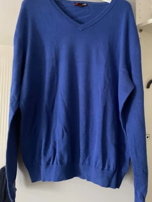 Kornblå stickad tröja med V-ringning - En enkel och stilren kornblå stickad tröja med V-ringad hals. Tröjan har långa ärmar och ribbade muddar vid ärmslut och nederkant. Perfekt att matcha med jeans eller finbyxor.