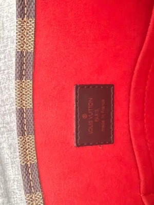 Louis Vuitton brun rutig handväska - Säljer en klassisk handväska från Louis Vuitton i brunt damier-mönster med insida i röd mocka. Väskan har flera innerfack och detaljer i mörkbrunt läder. Perfekt för dig som gillar lyxiga accessoarer och tidlös design.