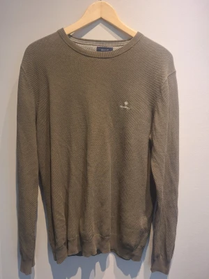 Olivgrön stickad tröja från GANT - Snygg olivgrön stickad tröja från GANT med rund halsringning och diskret logga broderad på bröstet. Tröjan har ribbade muddar vid ärmslut och nederkant. Perfekt för lager på lager och enkel att matcha med jeans eller chinos.