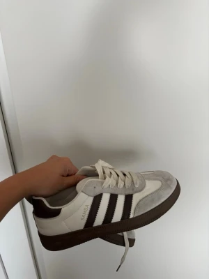 Adidas Samba sneakers vit/brun - Säljer ett par klassiska Adidas Samba sneakers i vitt och brunt med de ikoniska tre ränderna på sidan. Skorna har ovandel i mocka och skinn, samt snörning och brun yttersula. Perfekta för dig som gillar retrostil och streetwear.